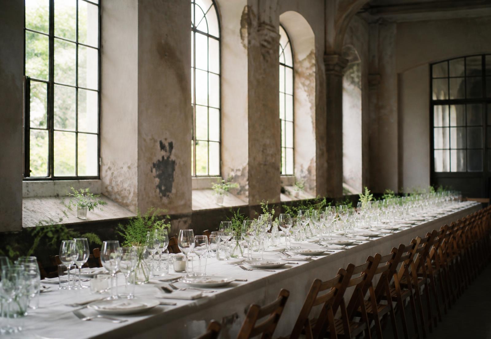 Long banquet table set with white linen in a light-filled hall with tall arched windows